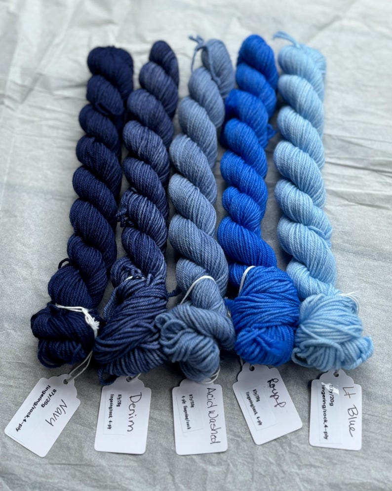 Pu&ograve; includere: Cinque gomitoli di filo blu in diverse tonalit&agrave; di blu, dal blu navy scuro al blu chiaro. Ogni gomitolo &egrave; etichettato con un nome e un peso: Navy, Denim, Acid Washed, Roupf, Lt Blue.