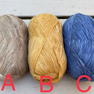 Puede incluir: Tres ovillos de hilo en diferentes colores: beige, amarillo y azul. El hilo es texturizado y tiene un aspecto r&uacute;stico.