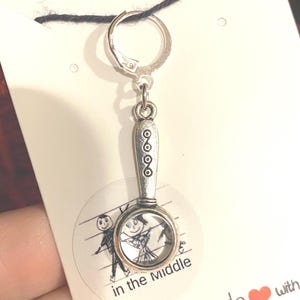 May include: A silver magnifying glass charm with a black and white drawing of two people inside the glass. The charm is attached to a silver jump ring and a black cord. The charm is on a white card with the text "in the middle" and "Handmade with love".