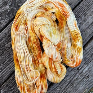 May include: A skein of white yarn with orange and yellow dye spots. The yarn is twisted and piled on a wooden surface.