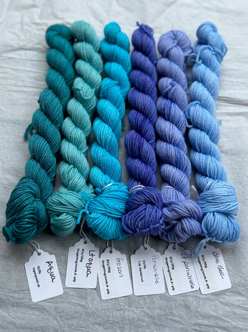 Pu&ograve; includere: Otto gomitoli di filato fingering in tonalit&agrave; di blu e turchese. Il filato &egrave; avvolto in gomitoli ed etichettato con i nomi "Blue Bell", "It Periwinkle", "Periwinkle", "Frozen", "Itagua" e "Aqua".