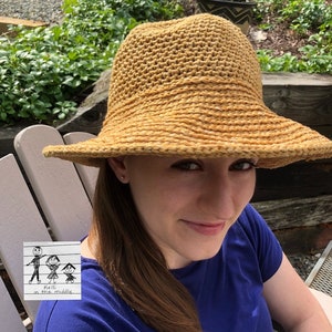 A person wearing a wide brimmed, crocheted, gold hat. The hat has a textured woven design. The person is wearing a blue shirt and has long brown hair.