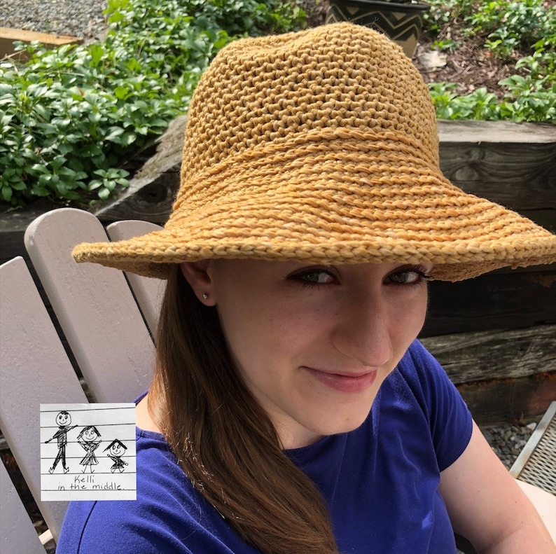A person wearing a wide brimmed, crocheted, gold hat. The hat has a textured woven design. The person is wearing a blue shirt and has long brown hair.