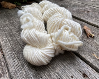 May include: Two skeins of white yarn, made of natural fibers, are shown on a wooden surface. The yarn is soft and fluffy, and it is perfect for knitting or crocheting.