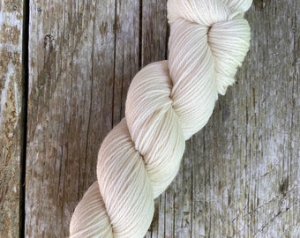 May include: A skein of off-white yarn, tightly wound and ready for knitting or crocheting. The yarn is a creamy color, and the texture appears soft and inviting. The yarn is resting on a weathered wooden surface.