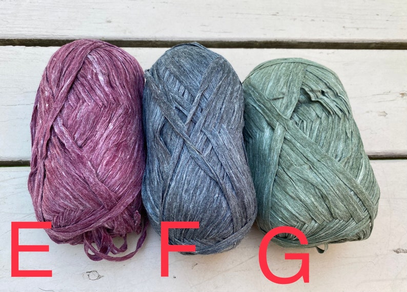 Puede incluir: Tres ovillos de hilo en diferentes colores: morado oscuro, gris oscuro y verde apagado. El hilo est&aacute; hecho de un material texturizado y arrugado.