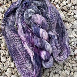 May include: A skein of hand-dyed yarn in shades of purple, lavender, and gray. The yarn is arranged in a loose, flowing pattern, with a twisted skein in the center. The yarn is likely made of wool or a similar fiber, perfect for knitting or crocheting projects.