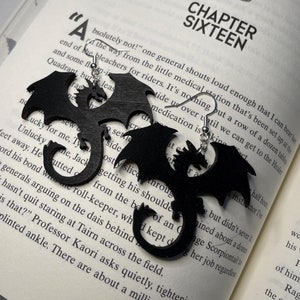 May include: A pair of black dragon earrings with a silver hook. The earrings are shown on a page of a book with the text "Chapter Sixteen" on the page.