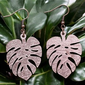 May include: A pair of earrings featuring a pink, metallic, leaf-shaped design. The earrings are made of wood and have a bronze-colored hook.