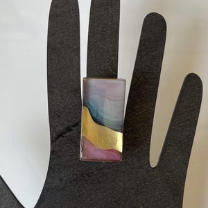May include: A rectangular ring with a colorful abstract design. The ring features a gold accent and shades of blue, pink, and purple. The ring is displayed on a black hand-shaped display.