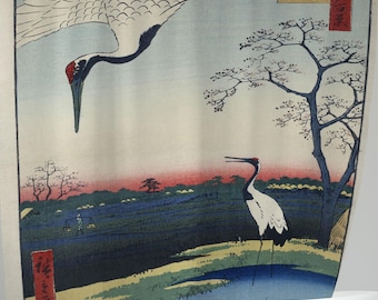 May include: A vintage Japanese woodblock print depicting two white cranes in a landscape setting. The cranes are flying and standing in a field with a river and trees. The print is in shades of blue, green, and red.