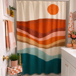May include: A shower curtain featuring a landscape print with a large orange sun above layered horizontal bands of rust, terracotta, peach, beige, light blue, and deep teal colors. The curtain hangs from a white rod with metal rings.
