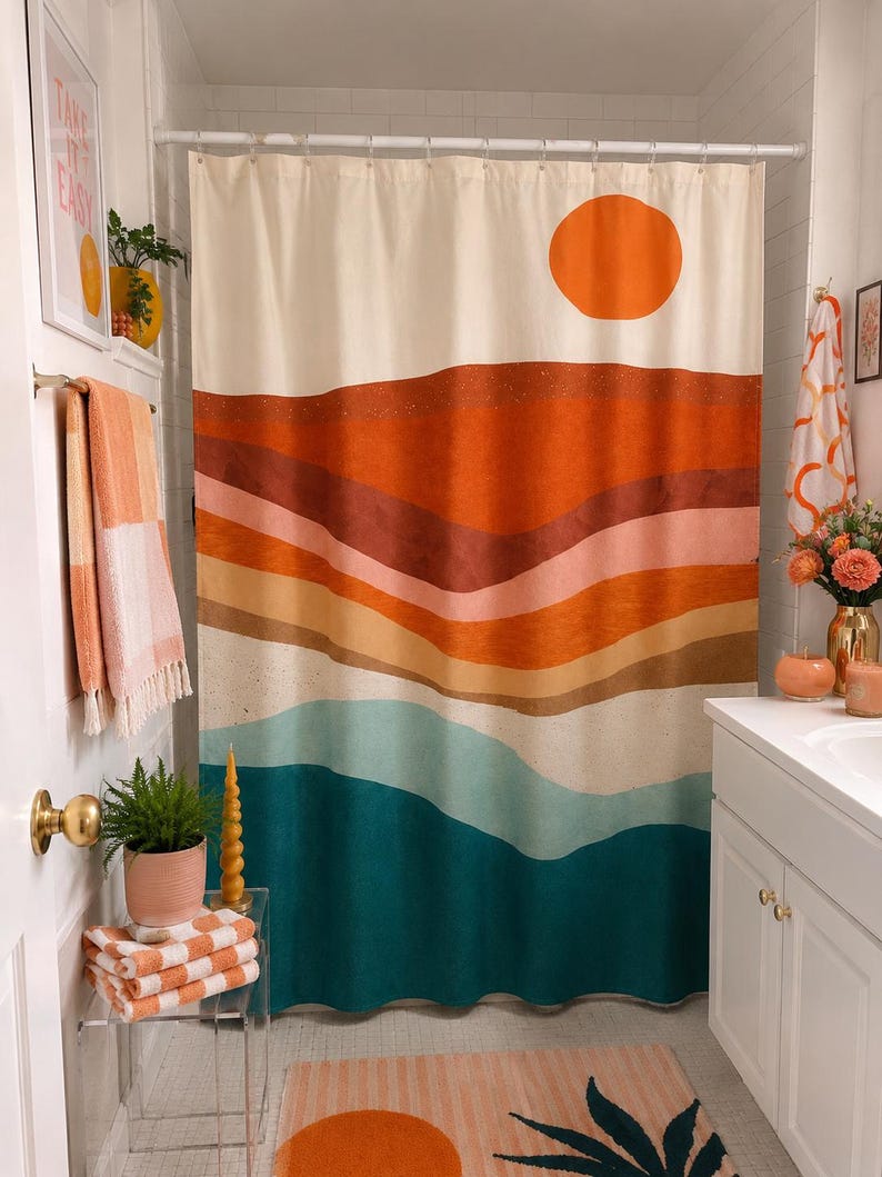 May include: A shower curtain featuring a landscape print with a large orange sun above layered horizontal bands of rust, terracotta, peach, beige, light blue, and deep teal colors. The curtain hangs from a white rod with metal rings.