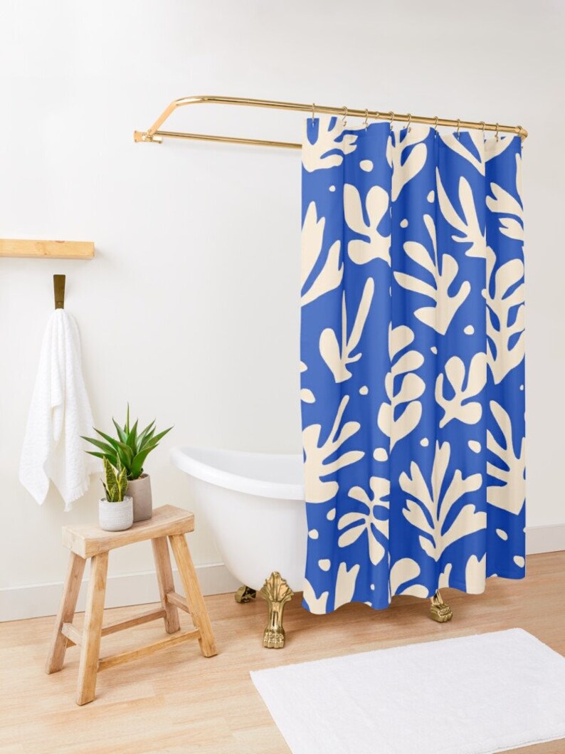 May include: A blue shower curtain with a cream leaf pattern hangs from a gold-coloured rod. The bathroom features a white bathtub with gold-coloured feet, a wooden stool, and a white towel.