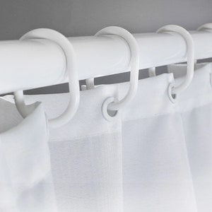 May include: A close-up shot of a white shower curtain rail with white, curved hooks supporting a white shower curtain. The curtain has a simple design, and the hooks are evenly spaced. The overall look is minimalist and modern, suitable for a bathroom.