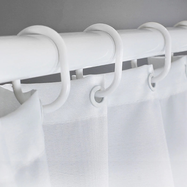 May include: A close-up shot of a white shower curtain rail with white, curved hooks supporting a white shower curtain. The curtain has a simple design, and the hooks are evenly spaced. The overall look is minimalist and modern, suitable for a bathroom.