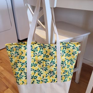 Peut inclure: Un sac fourre-tout jaune avec un motif de citrons et de feuilles. Le sac a un fond et des sangles de couleur crème. Il est accroché au dos d'une chaise blanche. L'arrière-plan comprend une machine à laver et un plancher en bois.