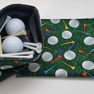 May include: A green fabric golf ball and tee holder with a black lining. The holder is filled with white golf balls and tees. The holder has a green fabric flap with a golf ball and tee pattern.