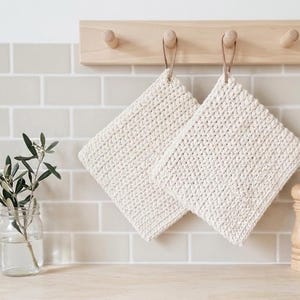 Könnte beinhalten: Zwei quadratische, cremeweiße gehäkelte Topflappen mit strukturiertem Maschenbild, jeweils mit einer kleinen braunen Lederschlaufe zum Aufhängen, befestigt an einer hellen Holzwandleiste mit vier Haken.