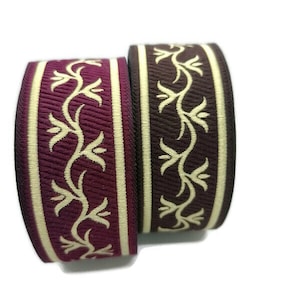 Könnte beinhalten: Zwei Spulen dekorativen Bandes. Eine ist ein tiefes Burgunderrot mit einem cremefarbenen Blumendesign und einem cremefarbenen Rand. Die andere ist ein dunkles Braun mit einem cremefarbenen Blumendesign und einem cremefarbenen Rand. Das Band wird wahrscheinlich für Bastelarbeiten oder Nähen verwendet.