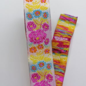 May include: A white ribbon with a colourful floral pattern. The ribbon features pink, yellow, orange, and blue flowers with green leaves. The ribbon is trimmed with a fringe of colourful threads.
