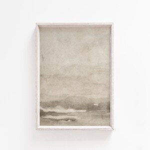 May include: A framed abstract art print featuring a watercolor-style landscape in muted beige and gray tones. The artwork is encased in a light-colored wooden frame, creating a minimalist aesthetic. The print's soft colors and textures evoke a sense of calm.