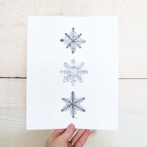 May include: Three watercolour snowflakes in shades of grey on a white background. The snowflakes are arranged vertically, with the largest snowflake at the top and the smallest snowflake at the bottom.