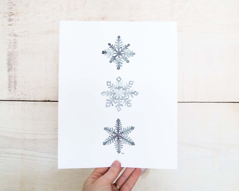 May include: Three watercolour snowflakes in shades of grey on a white background. The snowflakes are arranged vertically, with the largest snowflake at the top and the smallest snowflake at the bottom.