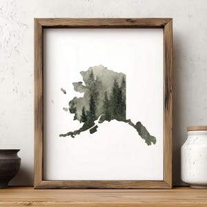 May include: A framed wall art print featuring the silhouette of the state of Alaska filled with a dark green forest landscape pattern. The artwork is enclosed in a rectangular wooden frame with a natural grain finish and a white mat border.