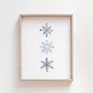 May include: A framed print featuring three watercolour snowflakes in shades of blue and grey on a white background.