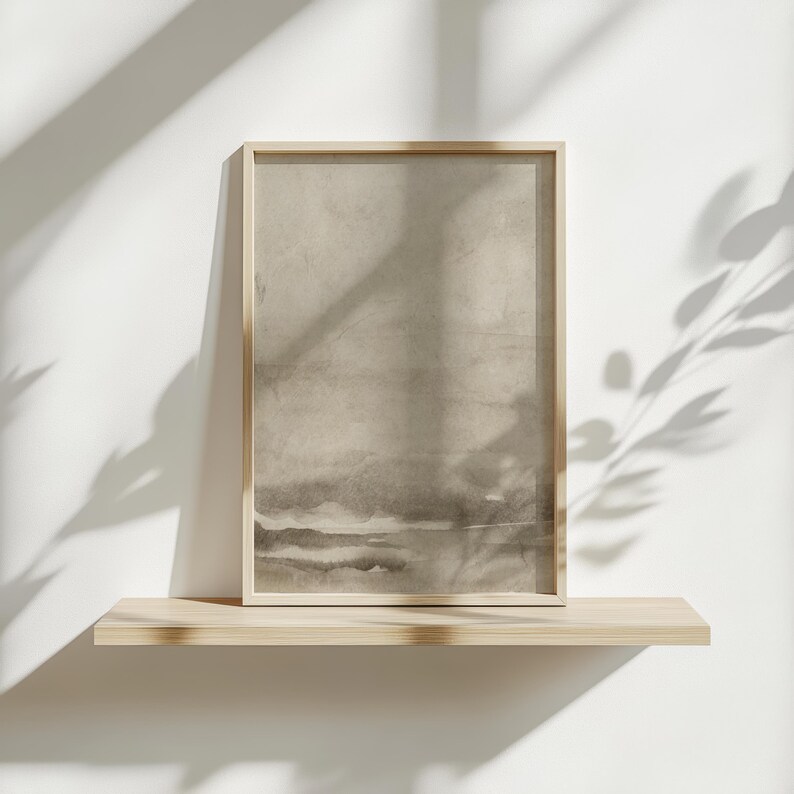 May include: A framed abstract art print featuring a neutral color palette of beige and gray tones. The artwork is displayed on a light wood shelf against a white wall, with shadows from foliage adding depth.