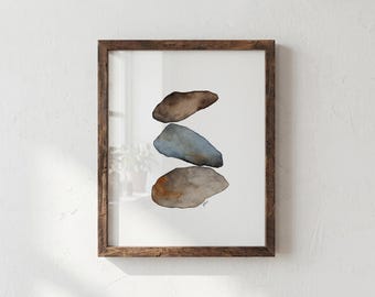 May include: A framed watercolor painting featuring three stacked, irregular stone shapes. The top stone is dark brown, the middle stone is a muted blue-gray, and the bottom stone is a larger, textured brown and tan shape. The frame is dark brown wood.