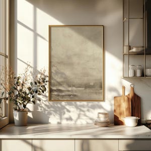 May include: A framed abstract landscape art print in a gold frame. The artwork features a muted color palette of beige, gray, and brown tones. The print is displayed in a kitchen setting, with a plant, cutting boards, and dishes visible.