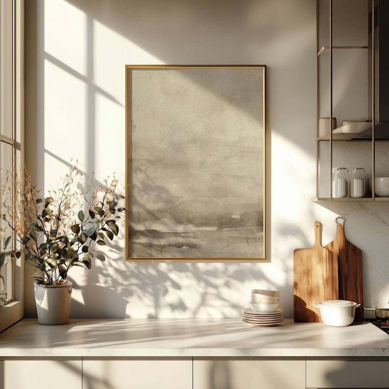 May include: A framed abstract landscape art print in a gold frame. The artwork features a muted color palette of beige, gray, and brown tones. The print is displayed in a kitchen setting, with a plant, cutting boards, and dishes visible.