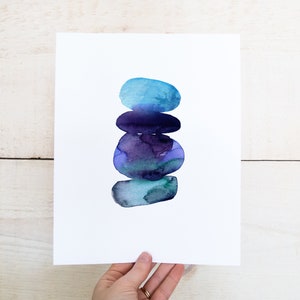 May include: A watercolor print featuring a stack of abstract, rounded shapes in shades of blue and purple. The artwork is displayed on a white background, held by a hand. The shapes resemble stacked stones or pebbles.