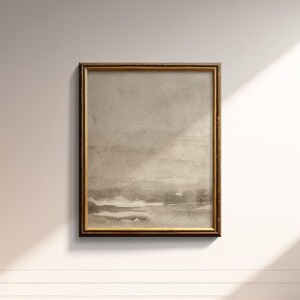 May include: A framed abstract landscape art print. The artwork features a muted color palette of beige, gray, and brown tones. The frame is a warm gold color. The print is rectangular and mounted on a white wall.