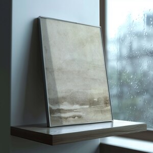 May include: A framed abstract art print with a silver metal frame. The artwork features a watercolor-style design in shades of beige and brown. The framed print is displayed on a wooden shelf next to a window with raindrops.