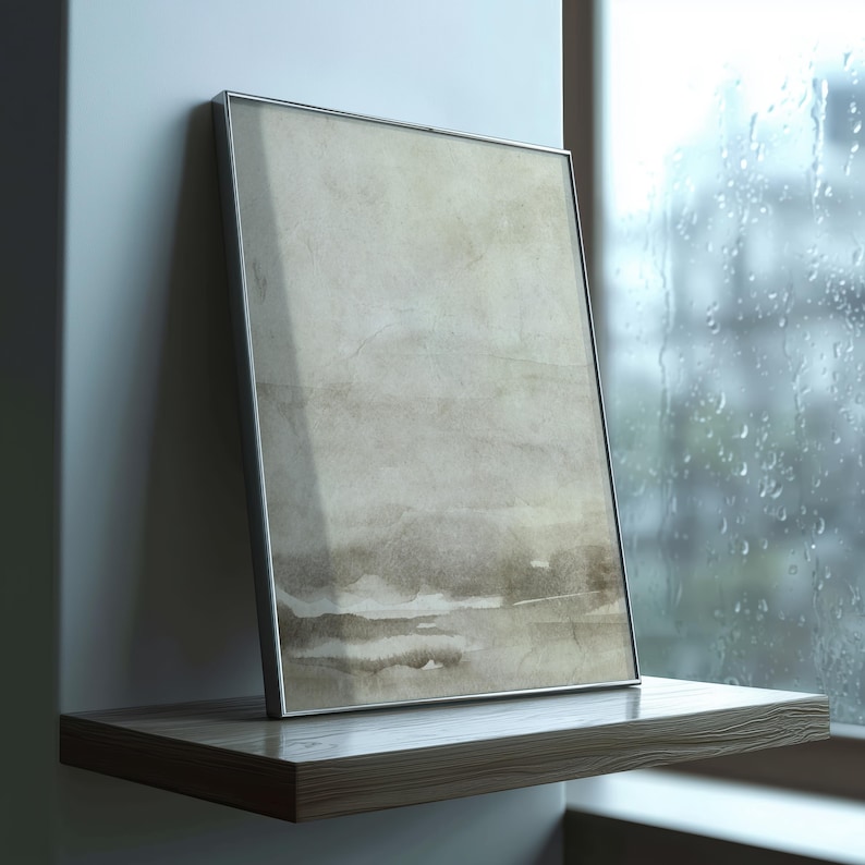 May include: A framed abstract art print with a silver metal frame. The artwork features a watercolor-style design in shades of beige and brown. The framed print is displayed on a wooden shelf next to a window with raindrops.