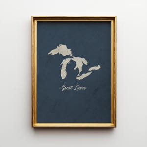 May include: A framed print featuring a map of the Great Lakes in a light beige color against a dark blue background. The words "Great Lakes" are written in a cursive font below the map. The frame is gold.