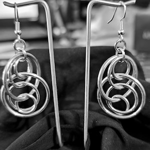 May include: A pair of silver earrings with a unique interlocking circle design. The earrings are hanging from a silver stand.