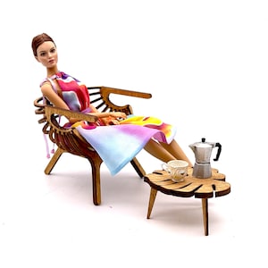 May include: A doll with brown hair wearing a sleeveless dress with a colorful abstract pattern of pink, orange, and blue. The doll sits in a brown wooden chair next to a small wooden table holding a miniature silver moka pot and a white cup.