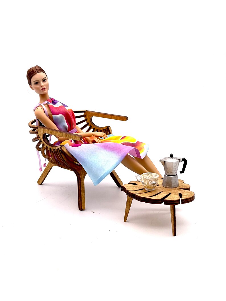 May include: A doll with brown hair wearing a sleeveless dress with a colorful abstract pattern of pink, orange, and blue. The doll sits in a brown wooden chair next to a small wooden table holding a miniature silver moka pot and a white cup.