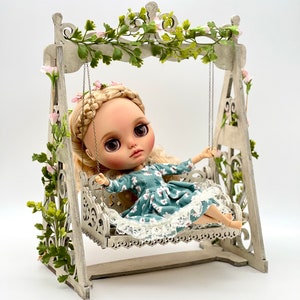 May include: A doll with blonde hair and a blue floral dress sits on a white wooden swing set decorated with green vines and pink flowers.