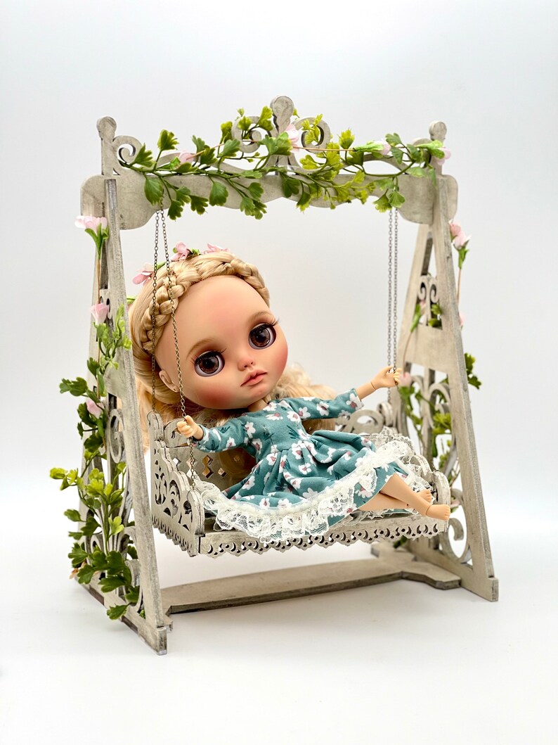 May include: A doll with blonde hair and a blue floral dress sits on a white wooden swing set decorated with green vines and pink flowers.