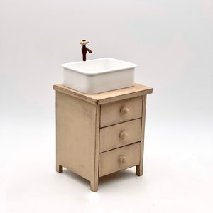 May include: A white porcelain sink with a brown faucet sits on top of a small wooden cabinet with three drawers. The cabinet is painted a light beige color and has a distressed finish.