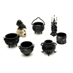 May include: A collection of miniature black Halloween-themed items including three cauldrons, one with the text Happy Halloween, a raven perched on a skull, a small candelabra, and a cylindrical container featuring a detailed skull pattern.
