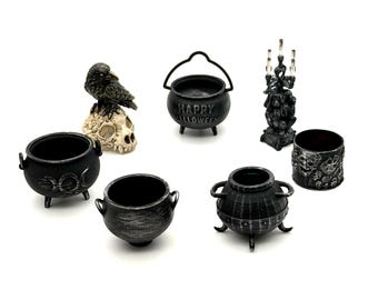 May include: A collection of miniature black Halloween-themed items including three cauldrons, one with the text Happy Halloween, a raven perched on a skull, a small candelabra, and a cylindrical container featuring a detailed skull pattern.