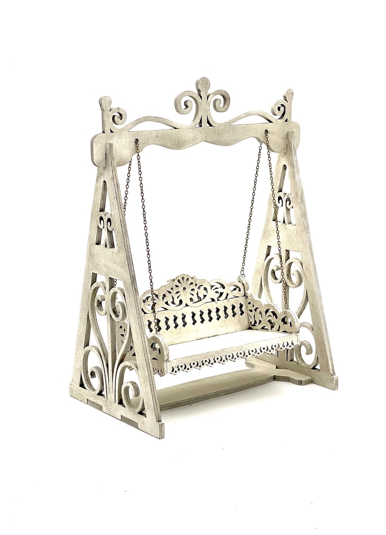 May include: White wooden swing set with intricate cutouts and a white cushioned seat. The swing is suspended from the frame by two chains.