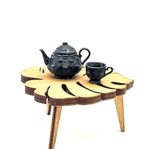 May include: A miniature black teapot and matching teacup with embossed heart patterns resting on a small, light brown wooden table shaped like a monstera leaf, featuring three angled legs and a layered plywood edge construction.