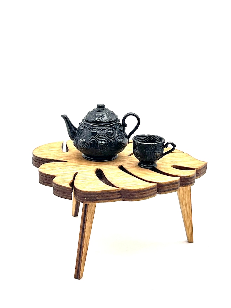 May include: A miniature black teapot and matching teacup with embossed heart patterns resting on a small, light brown wooden table shaped like a monstera leaf, featuring three angled legs and a layered plywood edge construction.